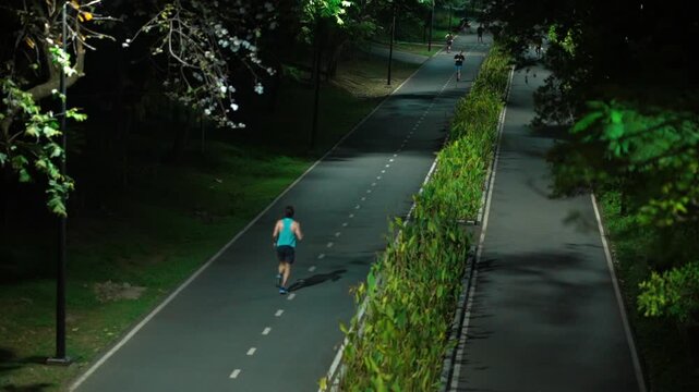 People running on park track at night with lights and trees, Joggers training outdoors on illuminated path in city park, evening fitness scene 4k