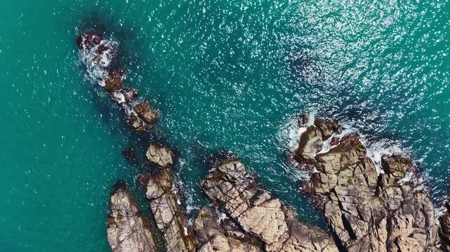Waves crash against dark rocks in clear blue water near a tropical beach. The sunlight reflects off the water surface creating bright patches. The scene is lively and natural.