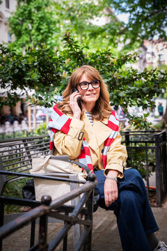 Smiling blond haired woman sitting on a city park bench with a smartphone and looking away while using a mobile phone outdoors