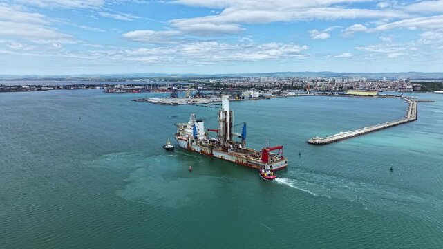 An industrial dredging vessel works in coastal waters close to an urban area. The vessel moves through the water as it performs dredging tasks under a clear sky.