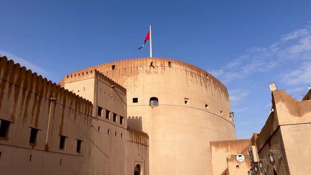 Historic Nizwa Fort after rain under clear blue sky with Omani flag and mosque minaret in background. Nizwa, Oman - May 14 2026