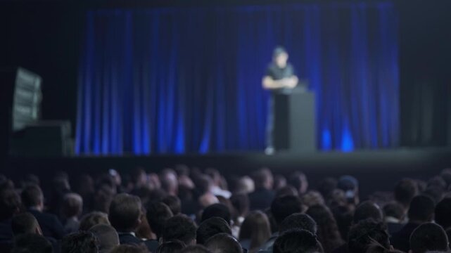 Large audience watching lone speaker deliver presentation on dimly lit stage, corporate conference atmosphere, focus on crowd and event.