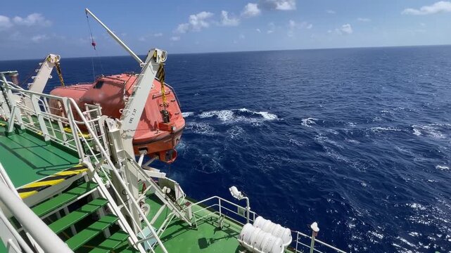 A bright orange fast rescue boat secured on its launching davit on the deck of a commercial vessel, with a background of choppy ocean waves during transit in windy maritime conditions