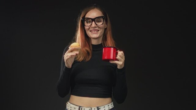 Woman holds red mug and cupcake with hands, wearing large glasses and black top, showing midriff and smiling in studio; cheerful warmth.