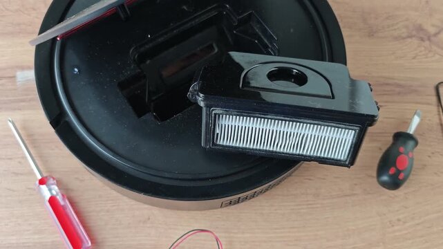 Robot vacuum cleaner repair process showing disassembly of the device, removal of the filter, and visible tools on a wooden surface in a well-lit indoor setting
