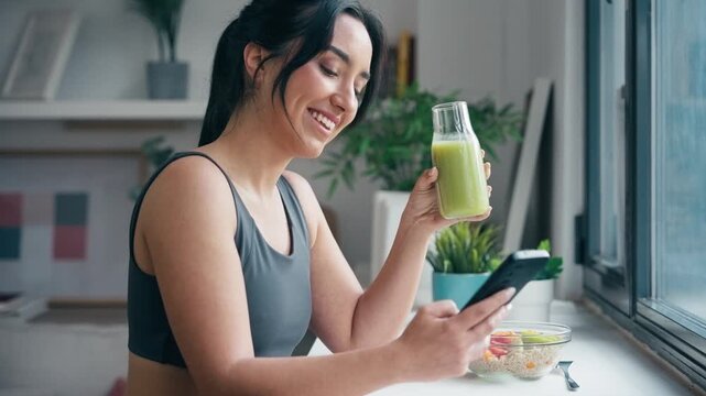 Video of fitness woman drinking a healthy green juice while using smartphone in the kitchen at home