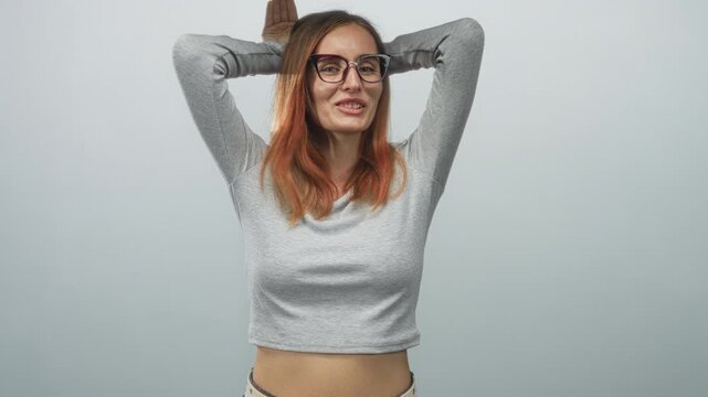 Woman with glasses and red hair placing hands on head, showing midriff in a neutral studio with soft lighting; playful confidence.