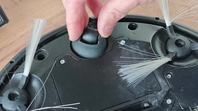 Hand adjusting components of a robot vacuum cleaner during repair, focusing on brush attachments and cleaning mechanisms on a wooden surface