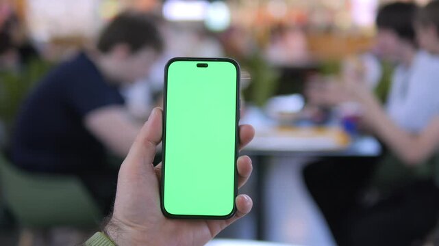 Hand holds phone with green screen. Background shows blurred cafe patrons. Focus remains on phone screen. No animals or plants visible. Scene suggests digital content display
