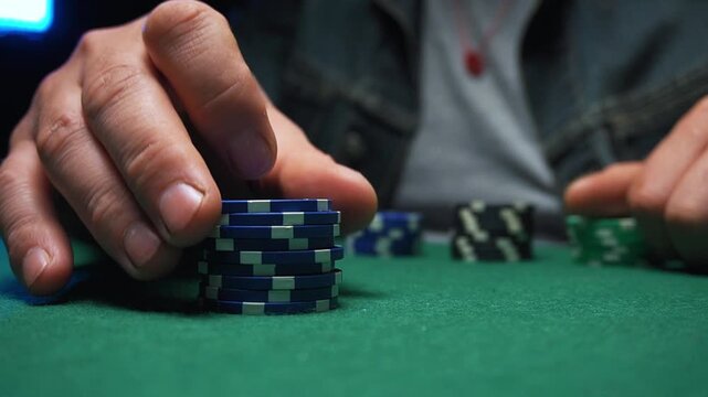 Close-up of male hand stacking blue poker chips on green felt table, with additional chips visible in the background during a card game