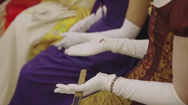 Close up of women in elegant historical dresses and white gloves. One holds a vintage fan, another an embroidered purse. Scene from a period drama, reenactment, or aristocratic ball