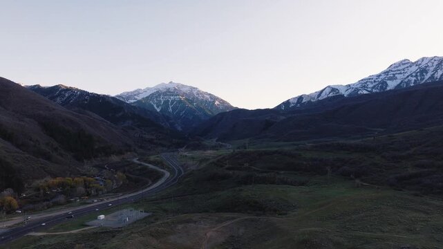Aerial view of Provo Canyon with a winding road through green hills and snow-capped mountain peaks at dusk Orem, Utah, United States.