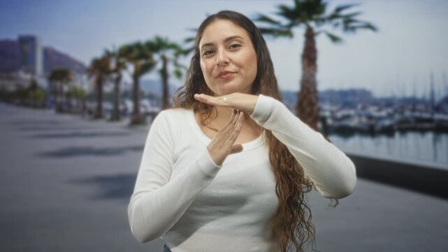 Woman brunette in white sweater making time out gesture with hands on a street by a marina with palm trees and boats visible; confidence.