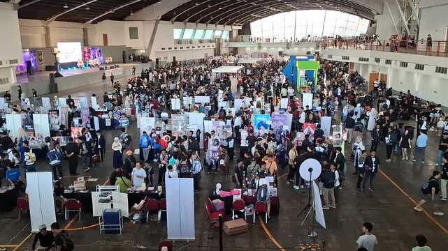 A high-angle with wide-shot video captures a cosplay convention inside a large convention hall, showcasing a vast crowd of attendees and numerous exhibition booths filling the expansive venue.