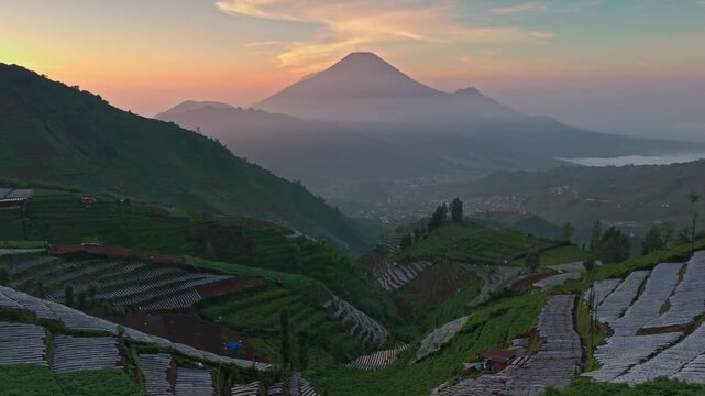 Aerial view of the terraced fields of Dieng Plateau with a misty valley and Mount Sindoro under a sunset sky in Kejajar, Central Java, Indonesia.