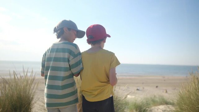 Best friends talking and sharing a moment of complicity and togetherness while looking at the sea from a sand dune on a sunny day during their summer vacation