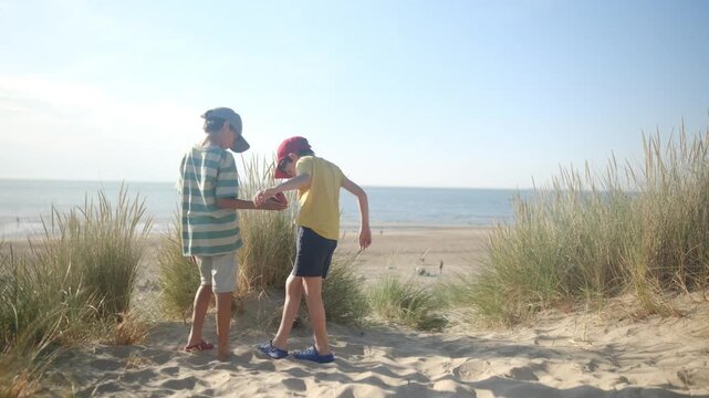 Two boys playing on a sand dune, emptying sand from a shoe while enjoying a sunny summer day at the beach, representing childhood friendship and carefree holidays