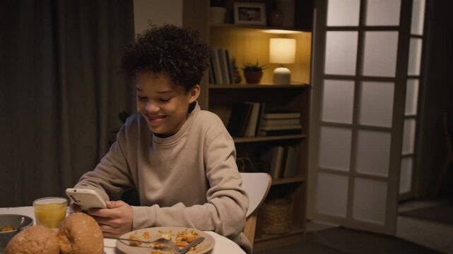 Medium shot of Black teenage boy reading messages online and using smartphone while sitting at dinner table at home, scene lit with warm evening lighting, copy space