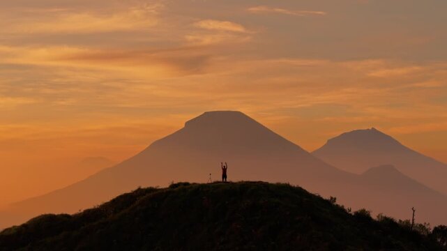 Aerial view of a person silhouette standing on a grassy hill overlooking the mountains of Dieng Plateau during a golden sunset Kejajar, Central Java, Indonesia.