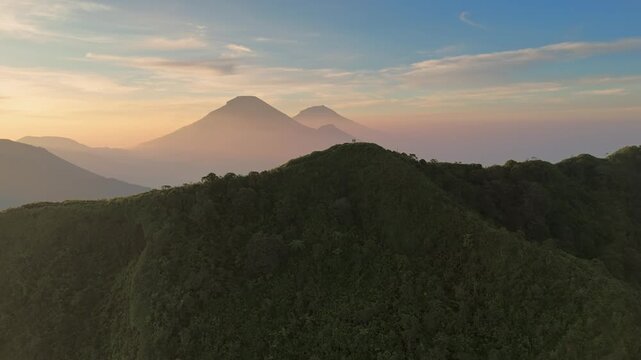 Aerial view of the lush green ridges of Dieng with volcanic peaks under a soft golden hour sky in Kejajar, Central Java, Indonesia.