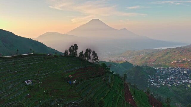 Aerial view of Dieng Plateau terraced agricultural fields on hillsides with Mount Sindoro in the misty background at sunrise Kejajar, Central Java, Indonesia.