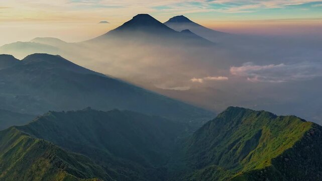 Aerial view of the Dieng Plateau volcanic mountains with green ridges and thick morning mist under a soft sunrise sky in Kejajar, Central Java, Indonesia.