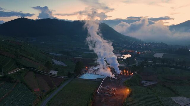 Aerial view of Dieng geothermal power plant with steam rising over agricultural terraces and mountains at dusk Kejajar, Central Java, Indonesia.