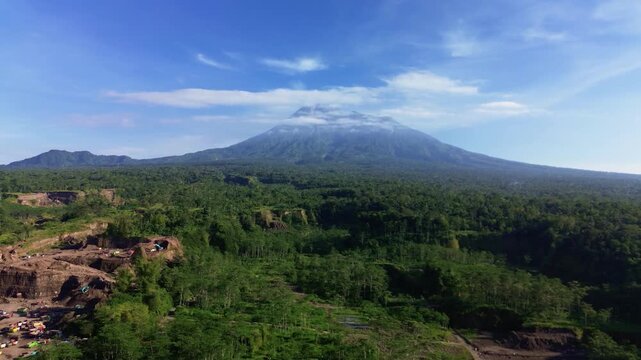 4K aerial footage of Mount Merapi with eroded cliffs and ravines caused by micro-mining in the foreground. A drone view shows green areas and eroded cliffs with the volcano in the background.