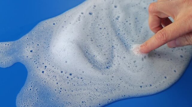 Close-up of a hand interacting with foamy soap on a blue surface, creating patterns and textures as the bubbles shift and reshape in a continuous flow of action
