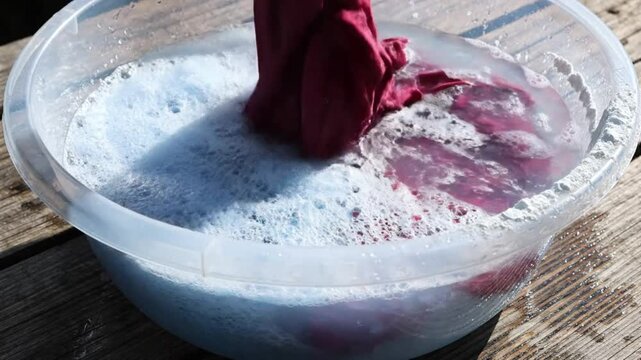 Washing colorful red fabric with blue soapy water in a clear plastic washbasin outdoors