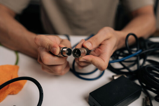 Audio technician hands holding xlr microphone cable, connecting male and female audio jacks, surrounded by tangled wires and various sound equipment on a white desk.