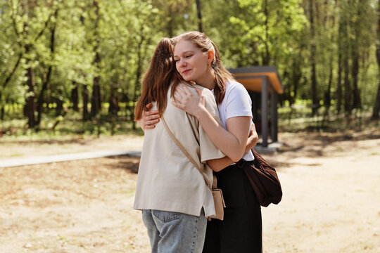 Two friends are hugging in a park filled with trees. They seem happy to see each other again. The sun shines down as they enjoy their time together outdoors