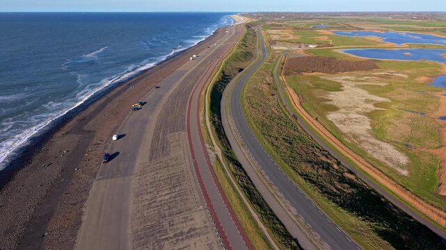 An aerial drone perspective stretches along the North Sea coastline, showing the asphalt sea dike infrastructure with roads and bike paths. The landscape contrasts the sea with inland wetlands.