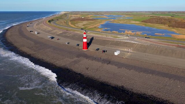 An aerial drone shot features the iconic red and white cast-iron Noorderhoofd lighthouse standing on the asphalt coastal dike. Waves crash on the shore, while camper vans park