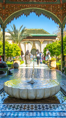 Fototapeta premium People walk through the Secret Garden in Marrakesh, Morocco. Sunlight shines down on the garden as visitors enjoy the lush plants and scenic views of architecture.