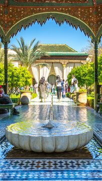 People walk through the Secret Garden in Marrakesh, Morocco. Sunlight shines down on the garden as visitors enjoy the lush plants and scenic views of architecture.