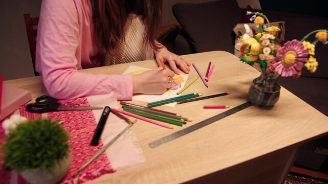 Woman sketching with colored pencils. Female caucasian artist relaxing in colorful drawing activity. Casual atmosphere of sketching nearby scissors, flowers in vase and colorful pencils on table