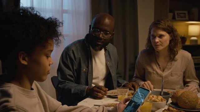Over shoulder shot of Black teenage boy using smartphone at dinner table and watching videos online ignoring parents and getting scolded