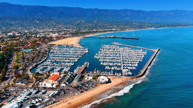 Foamy waves roll to the yacht club with multiple vessels. Iconic scenery of Santa Barbara, California, USA surrounded by hazy mountains. Aerial view.