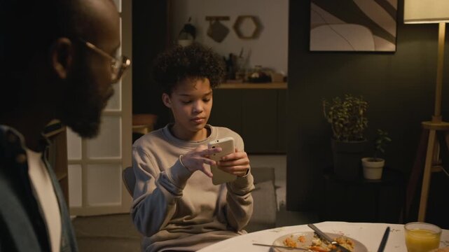 Medium shot of Black teenage boy fighting with mother scolding him for using smartphone at dinner table, candid scene lit by low evening lighting