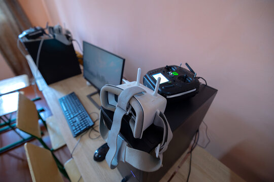 Educational and training equipment for FPV drone pilots including a virtual reality headset and a radio transmitter controller is set up on a desk with a computer monitor inside a room