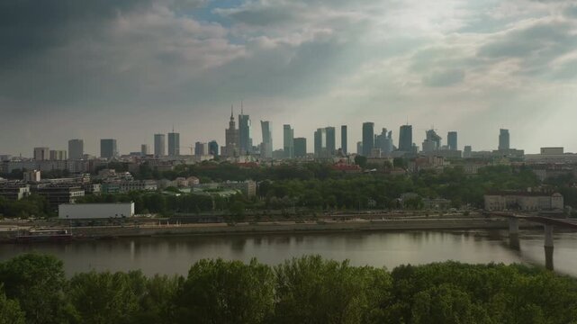Warsaw downtown skyline and business district skyscrapers stand beyond the Vistula river. The panorama pulls back over green parks, highway traffic and residential roofs under clouds.