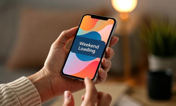 Hand holding smartphone displaying "Weekend Loading" on screen in cozy home setting