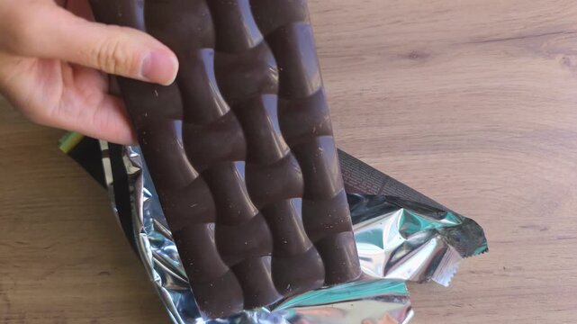 Hands unwrapping a chocolate bar from shiny foil packaging, revealing the smooth surface of the chocolate in a well-lit kitchen setting