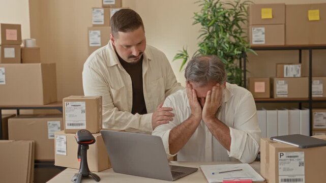 Two men packing parcels, older man covers face with hands while colleague places hand on shoulder in a building; stress support.