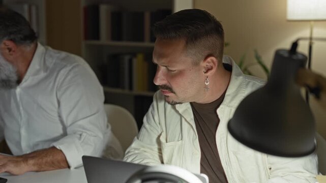 Two men typing on laptops with hands on keyboards and intent expressions in a building office at night; late concentration teamwork.