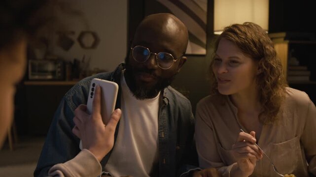 Over shoulder close up shot of Black teenage boy watching funny videos about school while using smartphone at dinner table and sharing meme with family, showing screen to smiling parents
