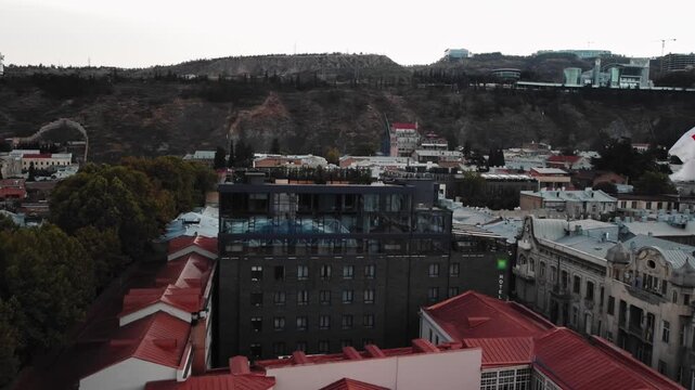 Aerial Tbilisi skyline with flag over historic rooftops and streets filmed by drone. Mix of modern and soviet buildings with distant caucasus hills on background under overcast sky. Travel theme
