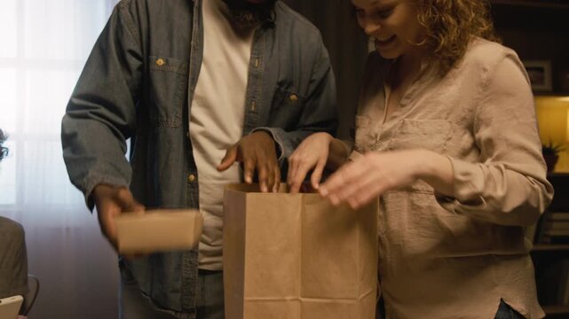Close up shot of multiracial family setting dinner table together and putting plates and takeout food out, cozy evening scene lit by low lighting, copy space