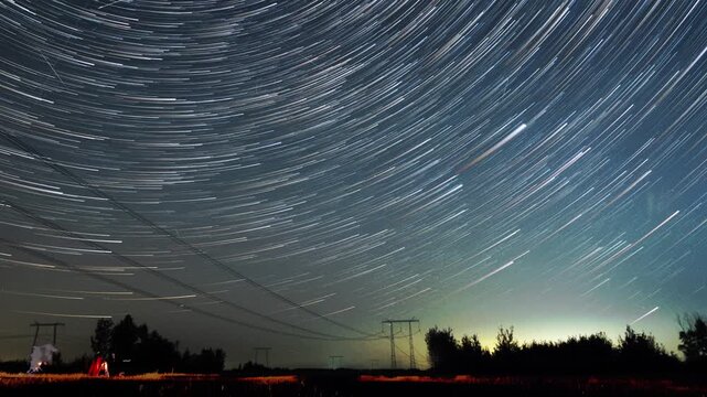 Wide star arcs over dark treeline, long exposure panorama with gentle Perseids streaks, transmission towers and distant lights on horizon, tranquil rural nightscape with cool tones and expansive sky.